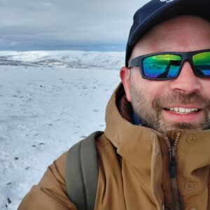 A person wearing a brown jacket, green sunglasses, and a navy cap stands outside in a snowy landscape. The background features snow-covered hills under a cloudy sky.