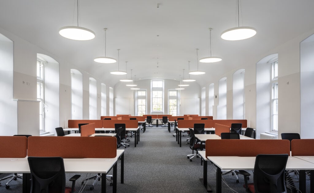 A spacious, modern office with high ceilings and large arched windows. The office is filled with rows of white desks, each equipped with black ergonomic chairs and orange privacy screens. Circular overhead lights provide bright, even illumination.
