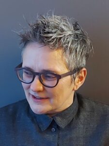 A person with short, gray hair and glasses smiles while looking slightly downward. They are wearing a dark buttoned-up shirt against a plain, dark background.