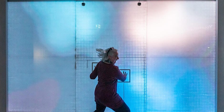A person with light-colored hair is enthusiastically running or dancing against a brightly lit, blue and purple checkered background with illuminated square patterns. The scene appears vibrant and dynamic.