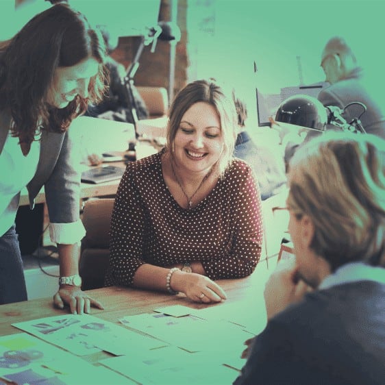 People meeting at a desk, looking at designs laid out on the table, green tint on image.