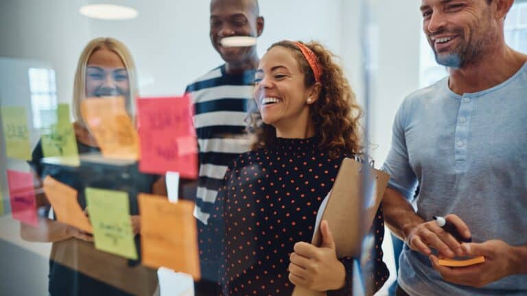 A group of four people stand smiling in front of a glass board covered with colorful sticky notes. One person holds a clipboard, while another holds a marker. The setting appears to be a collaborative workspace or office.