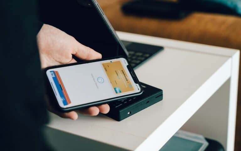 A person is holding a smartphone with an electronic payment app open, ready to make a contactless payment. The phone is near a payment terminal. A laptop is visible in the background on a white table.