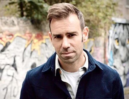 A man with short hair and a slight beard stands outdoors in front of a graffiti-covered wall. He wears a dark jacket over a white shirt, looking directly at the camera.