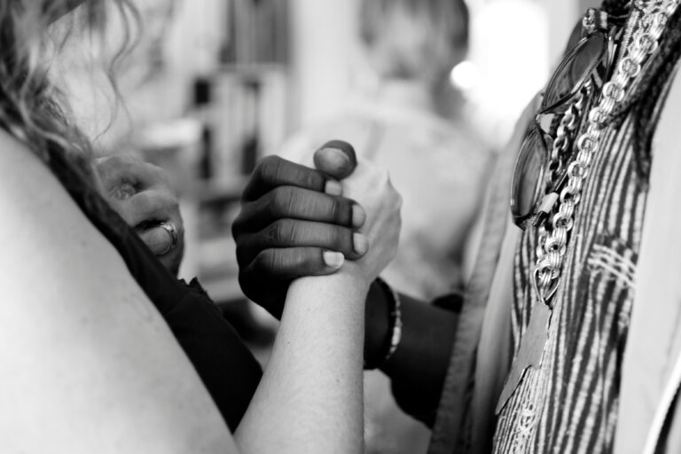 A black and white photo showing two people holding hands in a close-up. One person has light skin and the other has dark skin. The background is blurred, creating a focus on their clasped hands.