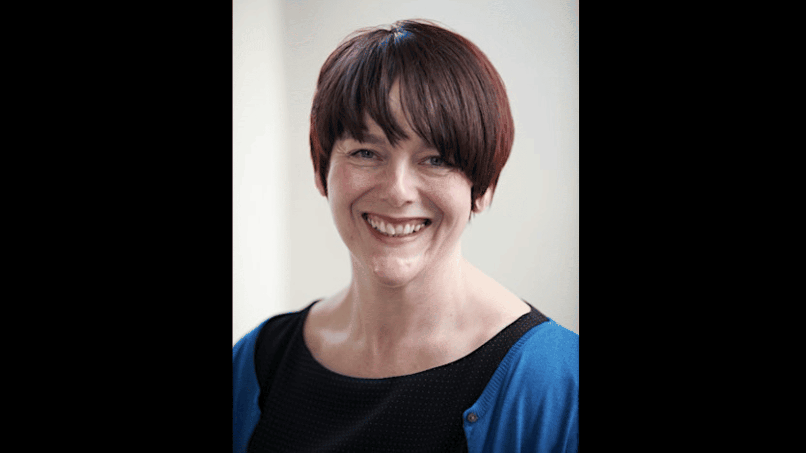 A person with short brown hair is smiling. They are wearing a black top with a blue cardigan against a neutral background.