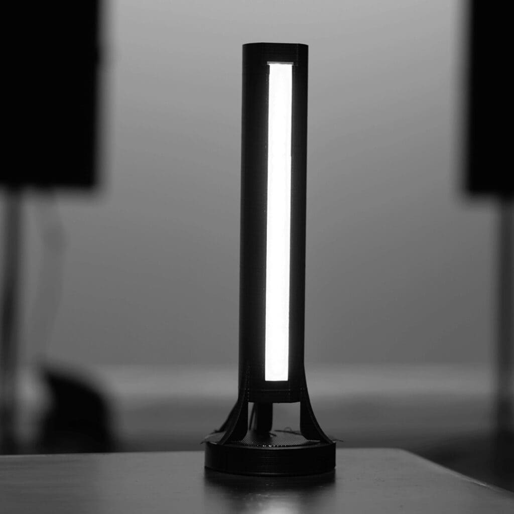 A modern, sleek cylindrical table lamp with a bright vertical light strip stands on a flat surface. The lamp is in front of a blurred background, creating a contrast between its illuminated design and the dark surroundings.