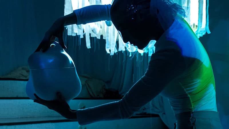A person in a dimly lit, blue-tinted room holds a large, abstract white sculpture. The background features a window with fringed white curtains, creating a dramatic contrast of shadows and light. The person's pose is dynamic and expressive.