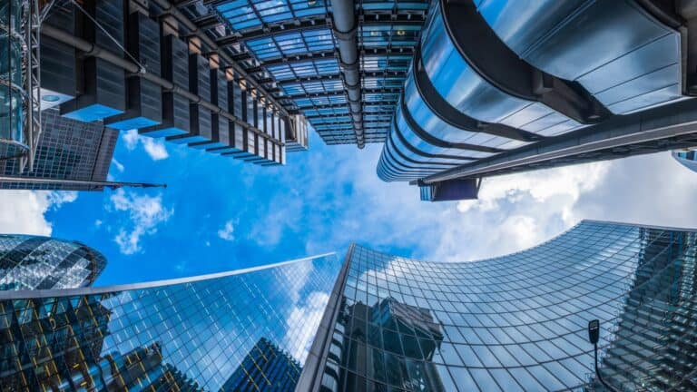 Upward view of modern skyscrapers with reflective glass and metal exteriors against a bright blue sky with white clouds. The tall buildings create a geometric pattern, converging towards the center of the image.