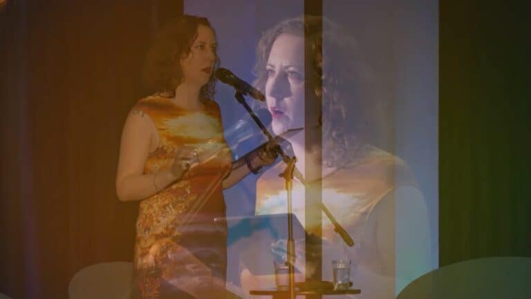 JL Williams performance: A woman in a colourful dress speaks into a microphone on stage. The image has an overlapping, transparent effect, showing her in duplicate. A glass of water sits on a table in front of her. The background is dimly lit.