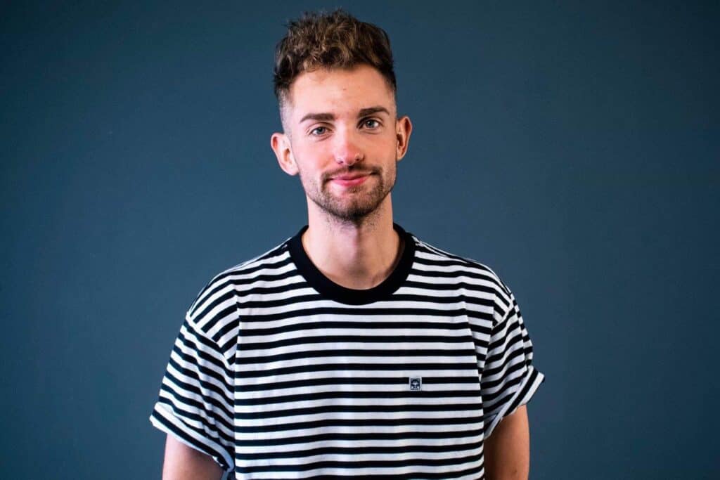 A man with short, curly hair and a beard is smiling. He is wearing a black and white striped T-shirt against a dark blue background.