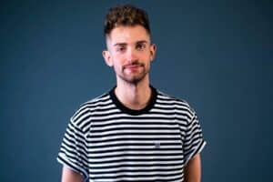A man with short, curly hair and a beard is smiling. He is wearing a black and white striped T-shirt against a dark blue background.