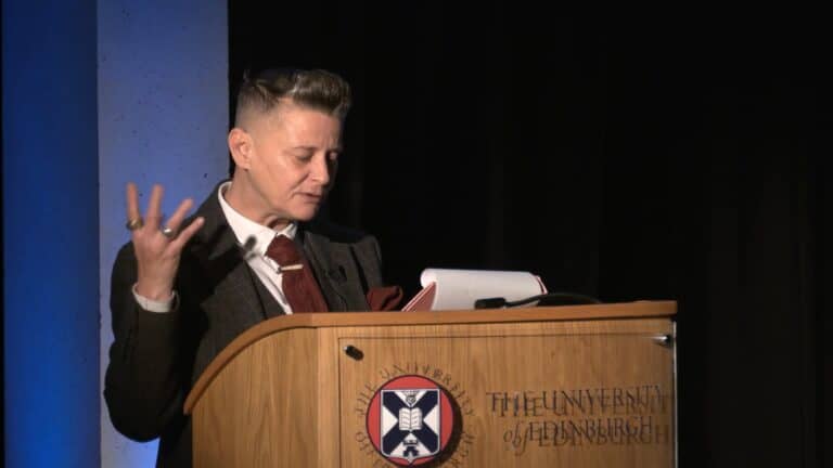 Joelle Taylor performance: A person with short hair speaks into a microphone while holding a book, standing at a podium with "The University of Edinburgh" on it. The background is dark with blue lighting on one side.