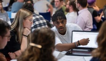 A group of people gathered around a table. One person is pointing at a graph on a laptop screen, engaging in discussion. Others are listening attentively. The setting appears to be a classroom or workshop.