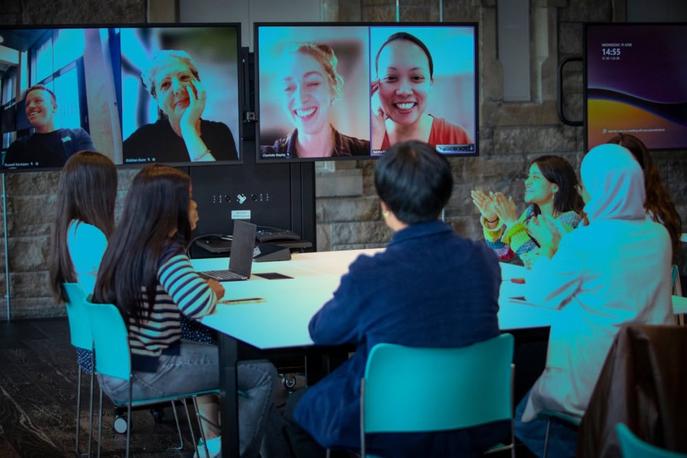 A group of people sit around a table in a modern meeting room, collaborating with three colleagues on video call shown on large screens. Some participants are smiling and clapping, suggesting a positive atmosphere.