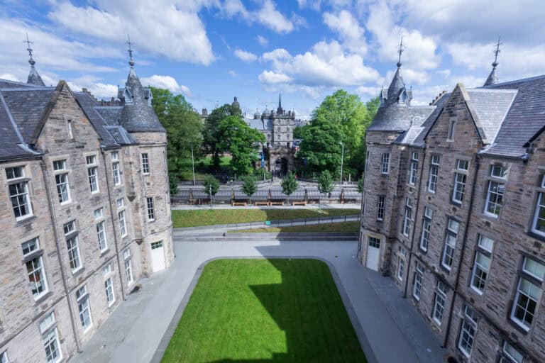 View through two historic stone buildings, framing a green lawn and a wide path, with a tree-lined road and a gothic-style gatehouse in the background under a blue sky with scattered clouds.